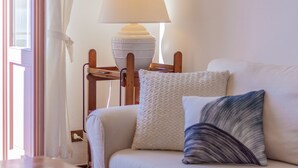 Ein weißes Sofa mit gemusterten Kissen, eine weiße Lampe auf einem Holztisch und ein Fenster mit weißen Vorhängen.