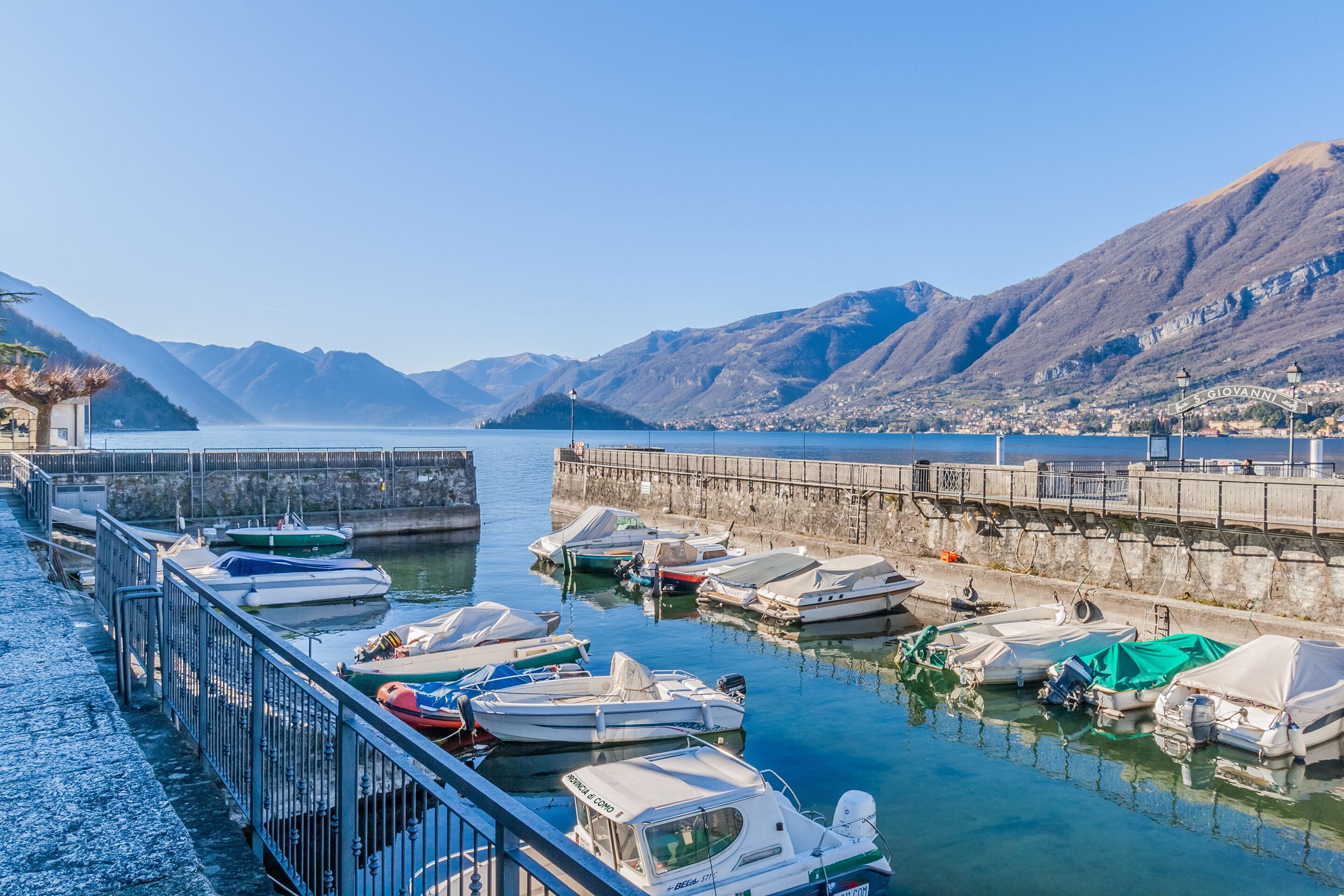 Uma marina com barcos atracados, um muro de pedra e montanhas ao fundo.
