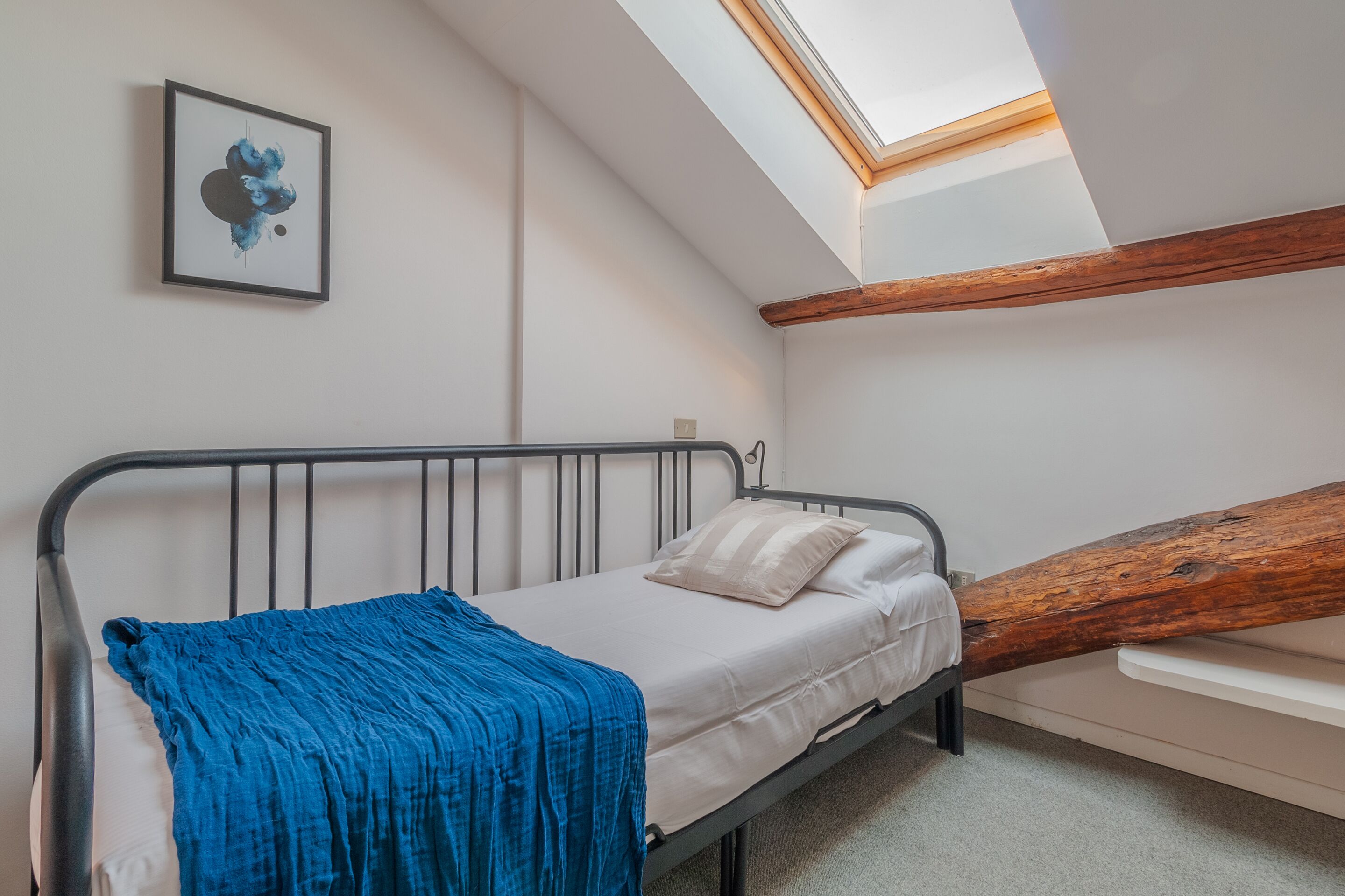 Un lit simple avec une couverture bleue et des oreillers blancs, une lucarne et une poutre en bois.