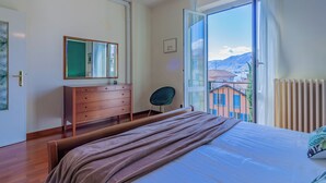 Une chambre à coucher comprenant un lit, une commode, une chaise et une grande fenêtre donnant sur des immeubles et des montagnes.