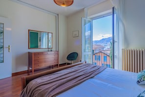 Une chambre à coucher comprenant un lit, une commode, une chaise et une grande fenêtre donnant sur des immeubles et des montagnes.