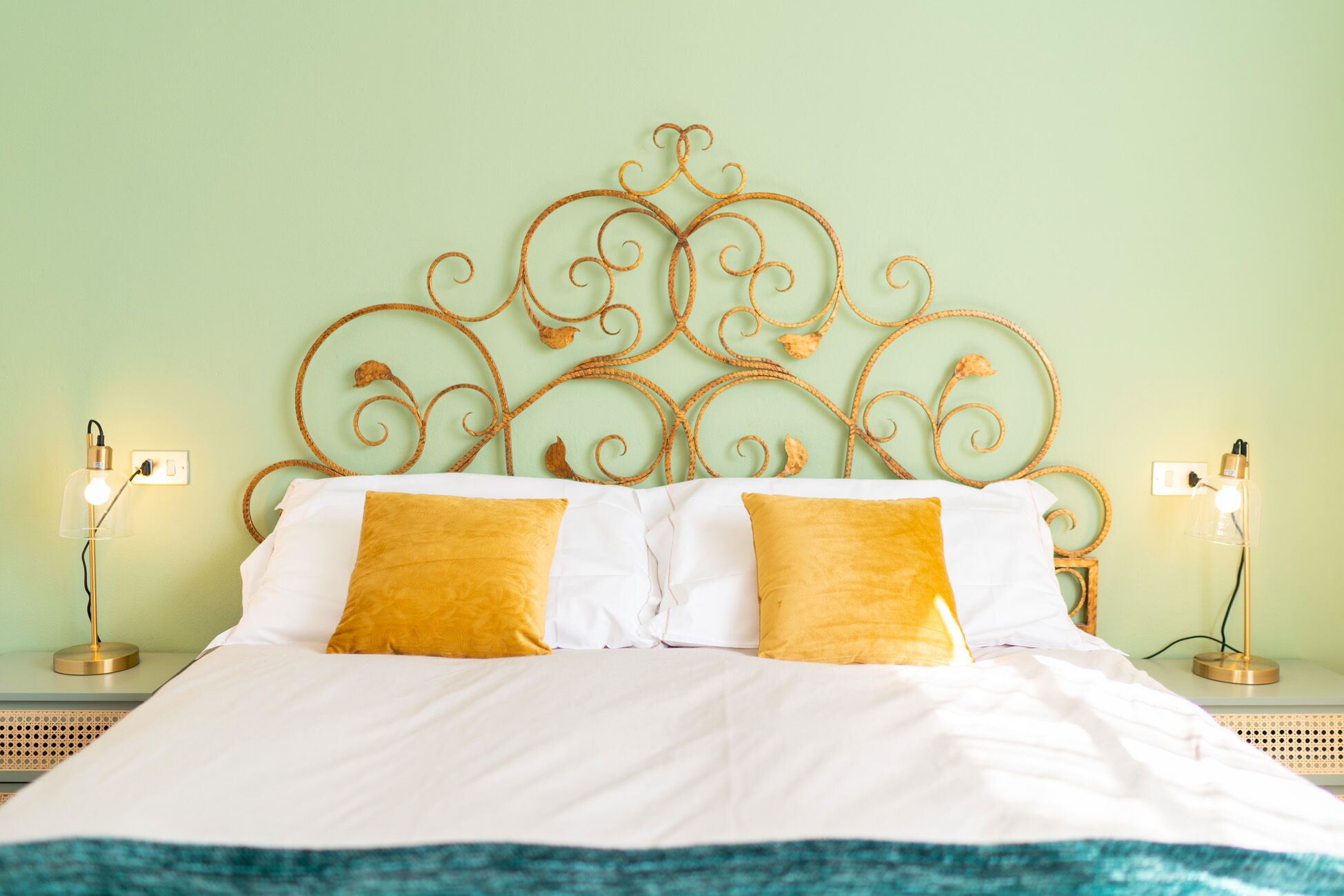 A neatly made bed with white linens and two mustard yellow pillows, a metal headboard with intricate designs, and two bedside lamps on matching nightstands.