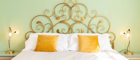 A neatly made bed with white linens and two mustard yellow pillows, a metal headboard with intricate designs, and two bedside lamps on matching nightstands.