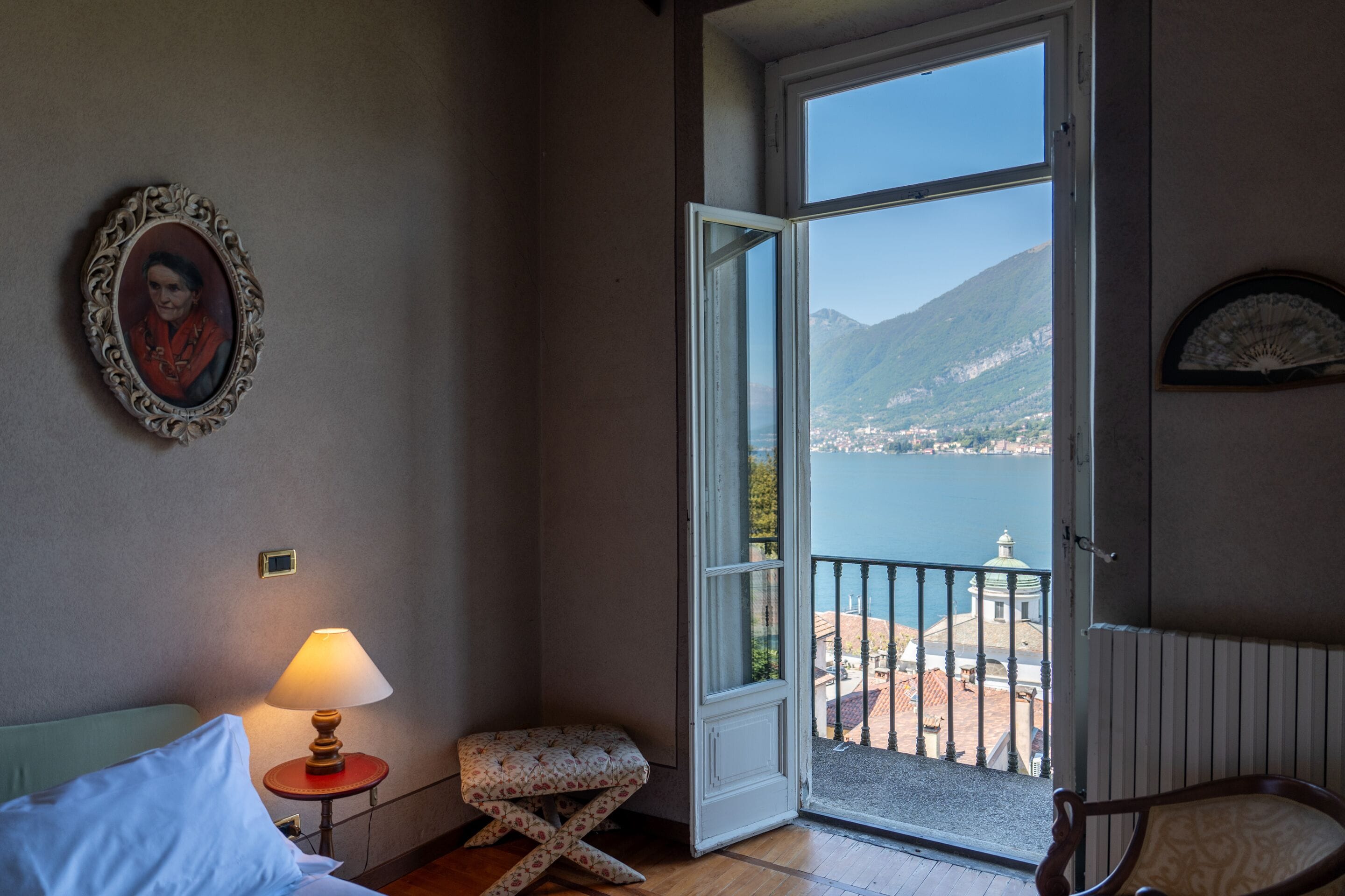 Ein Zimmer mit einem Bett, einer Nachttischlampe, einem kleinen Tisch, einem gemusterten Hocker und einer Tür zu einem Balkon mit Blick auf ein Gewässer und Berge.