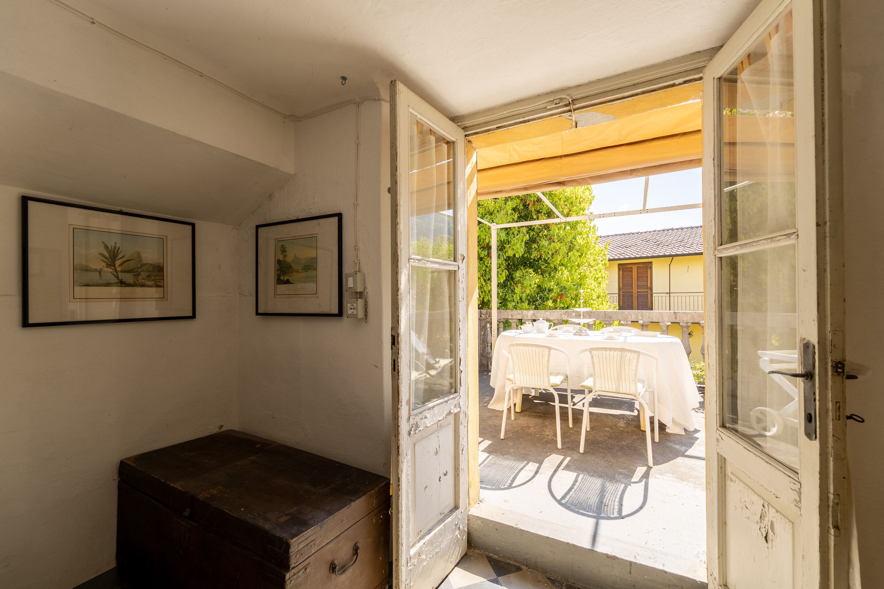 An open door leading to an outdoor patio with a table and chairs, framed pictures on the wall, and a wooden chest.