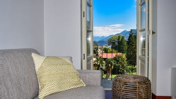A grey sofa with a patterned cushion and a wicker stool in a room with a view of mountains and greenery through open doors.