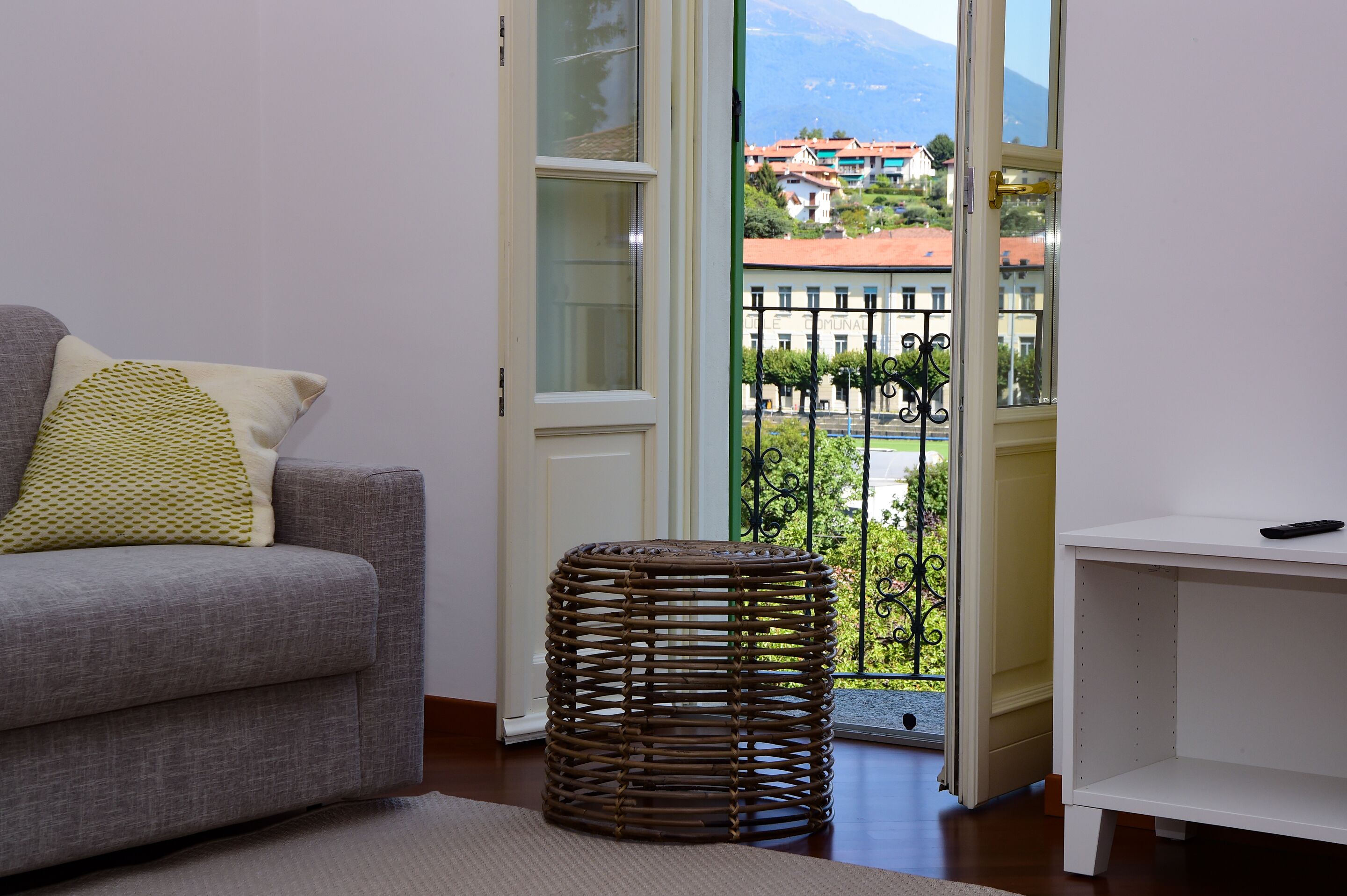 A living room with a grey sofa, a woven stool, and a view of a balcony with a railing and buildings in the background.