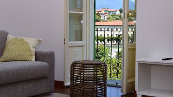 A living room with a grey sofa, a woven stool, and a view of a balcony with a railing and buildings in the background.