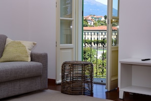 Una sala de estar con un sofá gris, un taburete tejido y vistas a un balcón con barandilla y edificios al fondo.