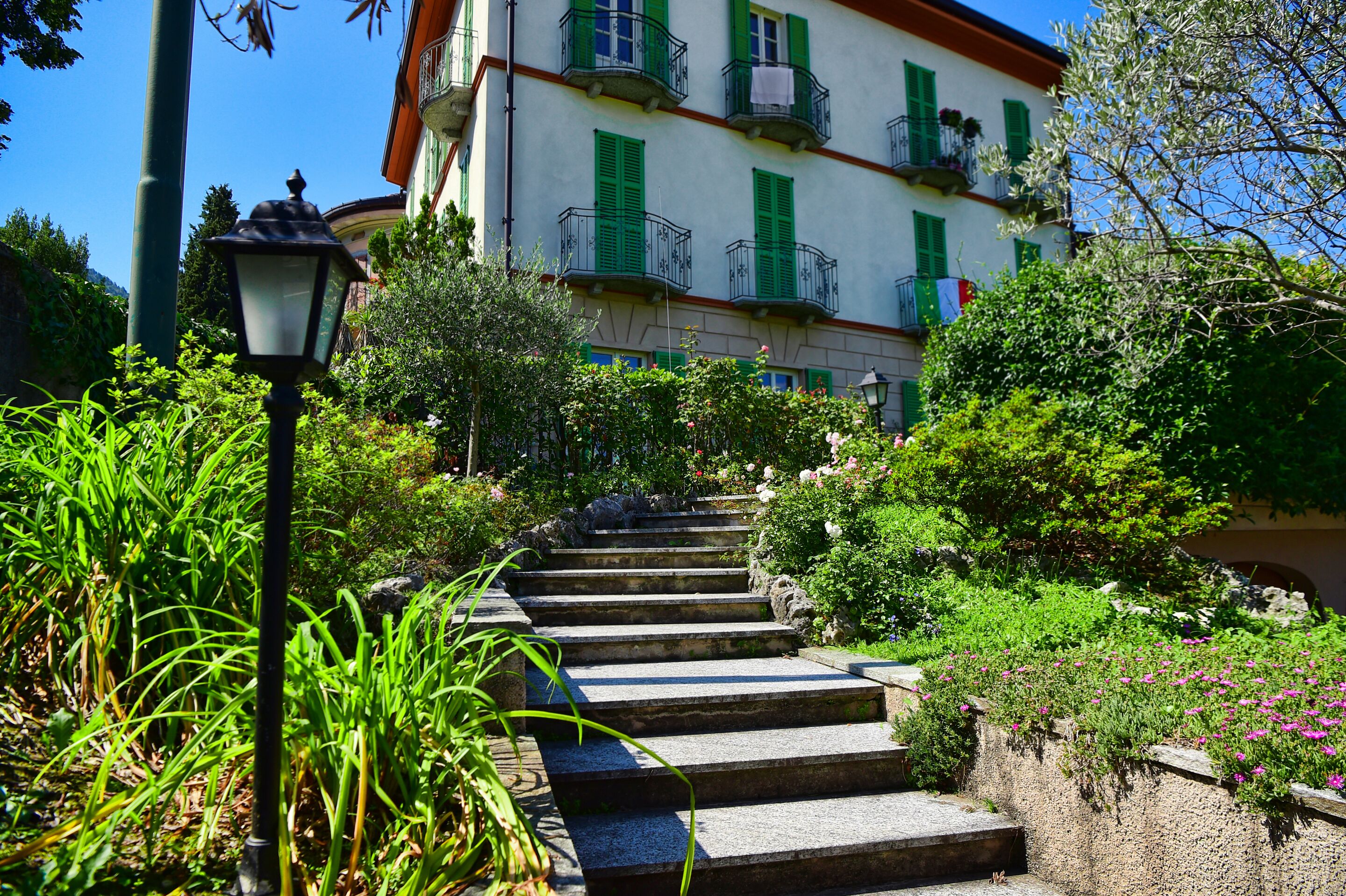 Degraus de pedra levam a um edifício com portaria verde e varandas, cercado por vegetação exuberante e um poste de luz.
