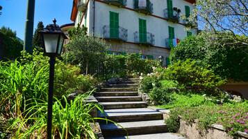 Degraus de pedra levam a um edifício com portaria verde e varandas, cercado por vegetação exuberante e um poste de luz.