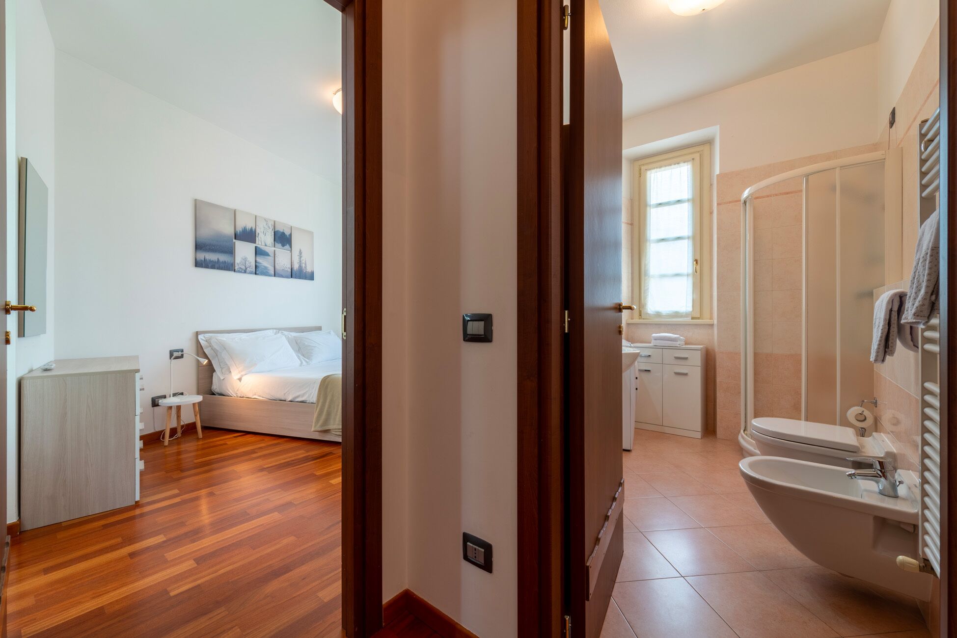 A hotel room with a wooden floor, a bed, a bedside table, a nightstand, a wall-mounted light, a window with curtains, a bathroom with a toilet, a bidet, and a towel rack.