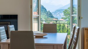 Un coin repas avec une table et des chaises, une vue sur un paysage montagneux par de grandes fenêtres ouvertes, et un balcon agrémenté d’une balustrade en fer forgé.