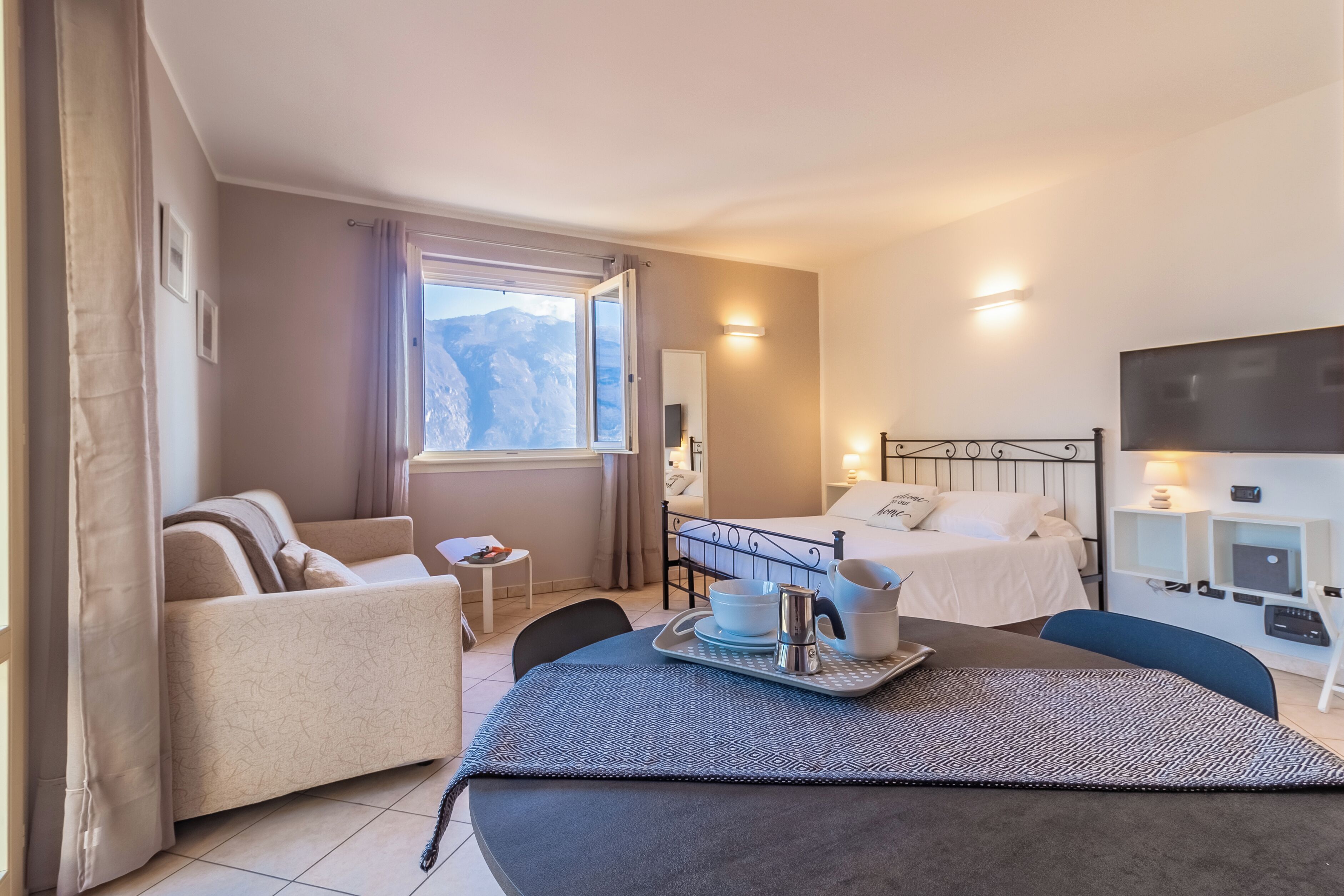 Habitación de hotel con cama, sofá, mesa de comedor con bandeja, televisor y ventana con vistas a la montaña.