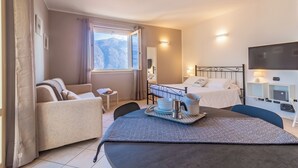 A hotel room with a bed, a sofa, a dining table with a tray, a TV, and a window with a mountain view. - Incanto Apartment - One Bedroom Apartment, Sleeps 2 (Bellagio)