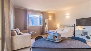 Habitación de hotel con cama, sofá, mesa de comedor con bandeja, televisor y ventana con vistas a la montaña.
