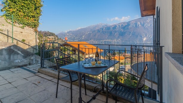 A balcony with a table set for two, a bottle of wine, and glasses. The balcony overlooks a mountainous landscape with buildings in the distance. - Incanto Apartment - One Bedroom Apartment, Sleeps 2 (Bellagio)