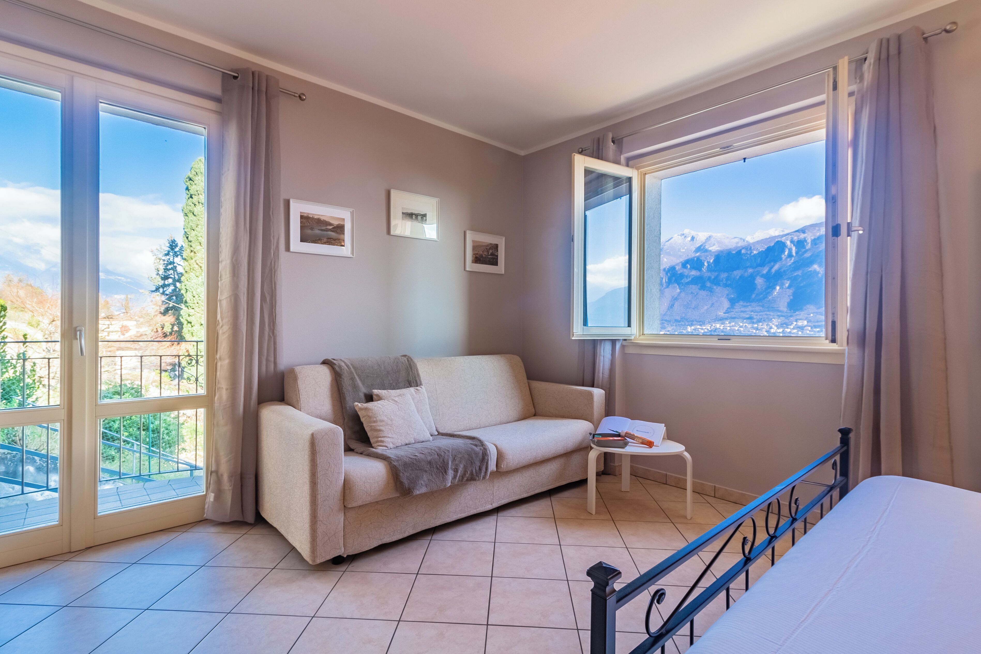 Una habitación con sofá, una mesita y una cama. Hay dos ventanas con cortinas, una de ellas con vistas a las montañas.