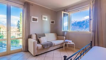Una habitación con sofá, una mesita y una cama. Hay dos ventanas con cortinas, una de ellas con vistas a las montañas.