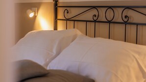 Ein ordentlich bezogenes Bett mit weißen Leinen und zwei Kissen, ein schwarzes Metall-Kopfende sowie eine an der Wand befestigte Leuchte.