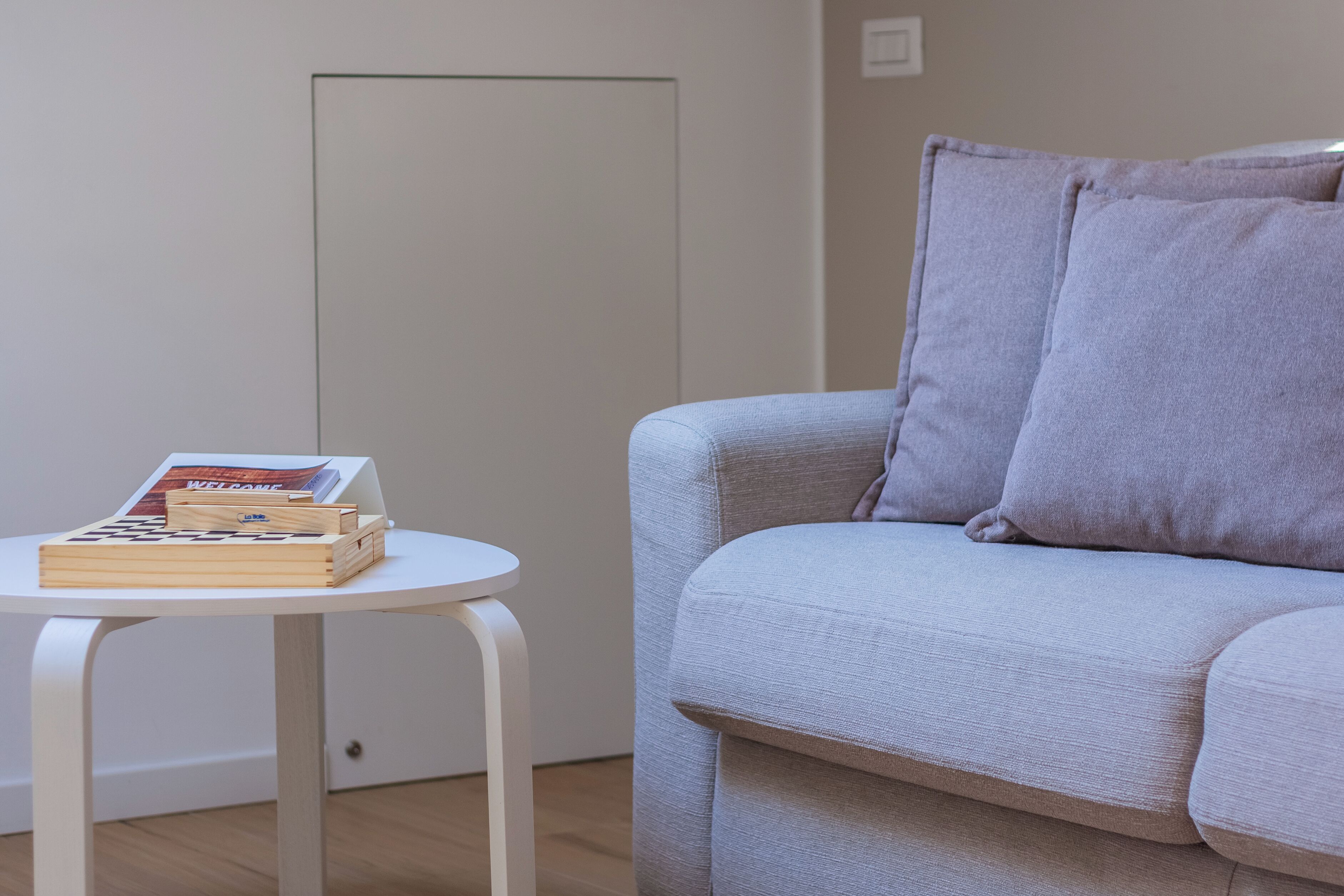 Una habitación con un sofá de gris claro, una pequeña mesa blanca con libros y una revista, y una puerta sencilla.
