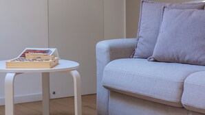 Ein Zimmer mit einem hellgrauen Sofa, einem kleinen weißen Tisch mit Büchern und einer Zeitschrift sowie einer schlichten Tür.