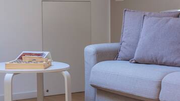 Una habitación con un sofá de gris claro, una pequeña mesa blanca con libros y una revista, y una puerta sencilla.