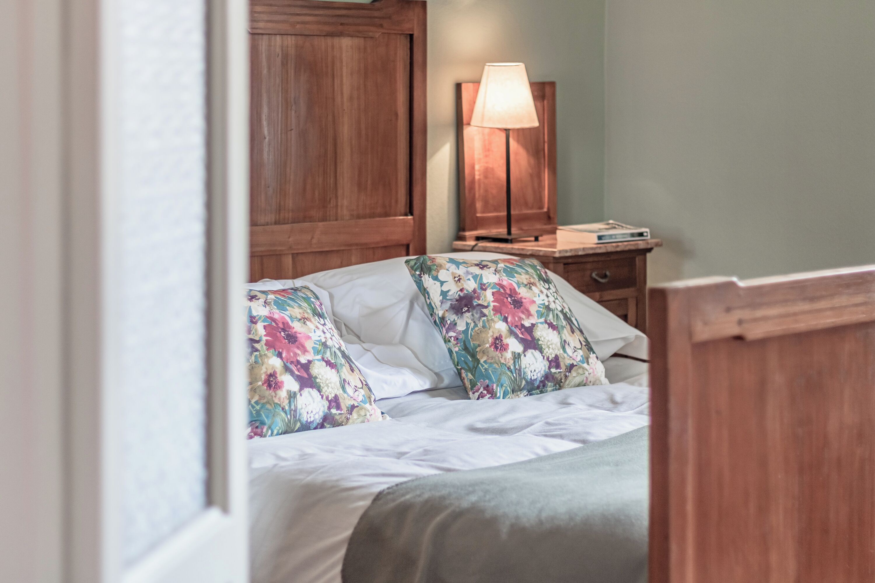 Un dormitorio con cabecera de madera, una mesita de noche con lámpara y almohadas estampadas con motivos florales.