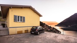 Ein gelbes Gebäude mit einem geparkten Auto davor, einem Metallgeländer und Blick auf ein Gewässer sowie Hügel im Hintergrund.