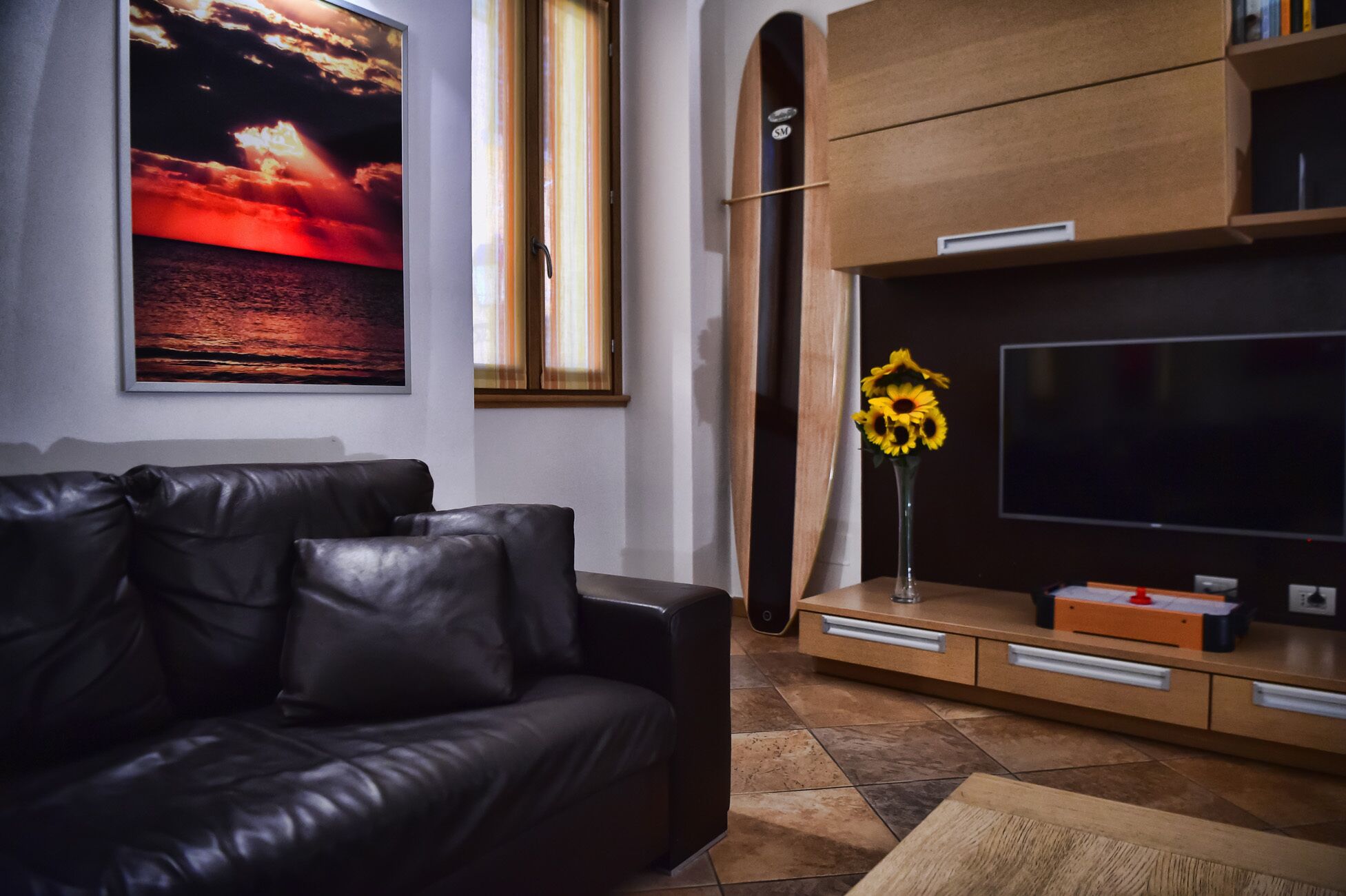 Een moderne woonkamer met een leren bank, een grote flatscreen-tv, een houten kast en een schilderij van een zonsondergang in een lijst.
