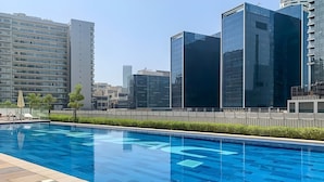 A swimming pool with a tiled deck, surrounded by modern buildings and a clear sky. - Elegant Reva 1BR - Pool & Gym (Dubai)