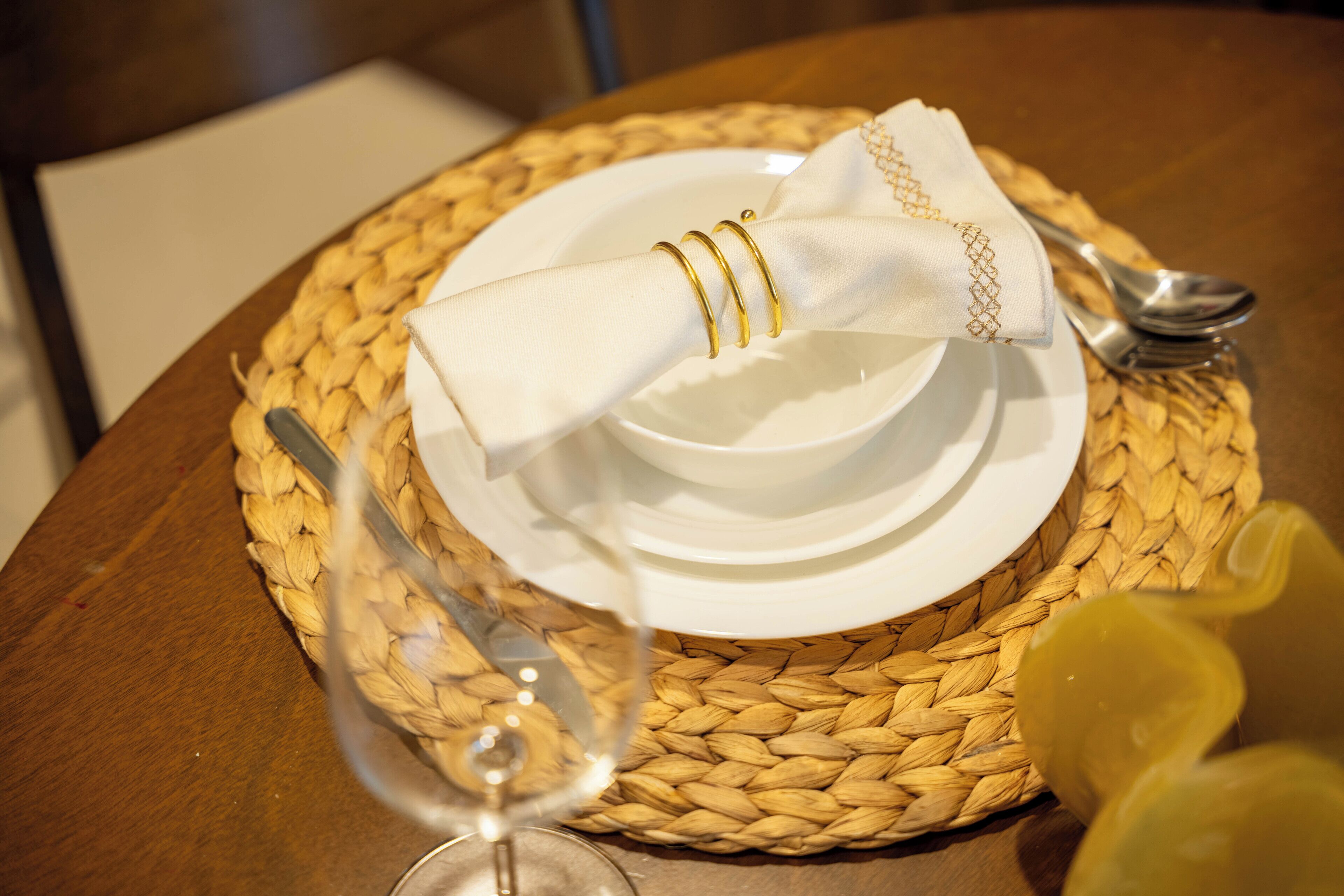 A table set with white plates, gold-rimmed napkins, and silverware on a woven placemat.