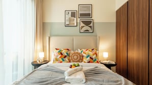 A neatly made bed with a towel, pillows, and a fruit bowl. There are two bedside tables with lamps and a wall with framed pictures. - Elegant Reva 1BR - Pool & Gym (Dubai)