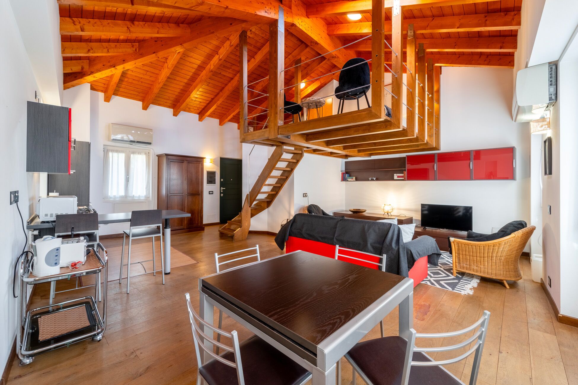 A spacious loft with a high ceiling, wooden beams, a dining area with a table and chairs, a living area with a sofa and TV, and a staircase leading to an upper level.