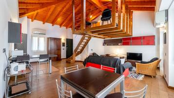 A spacious loft with a high ceiling, wooden beams, a dining area with a table and chairs, a living area with a sofa and TV, and a staircase leading to an upper level.