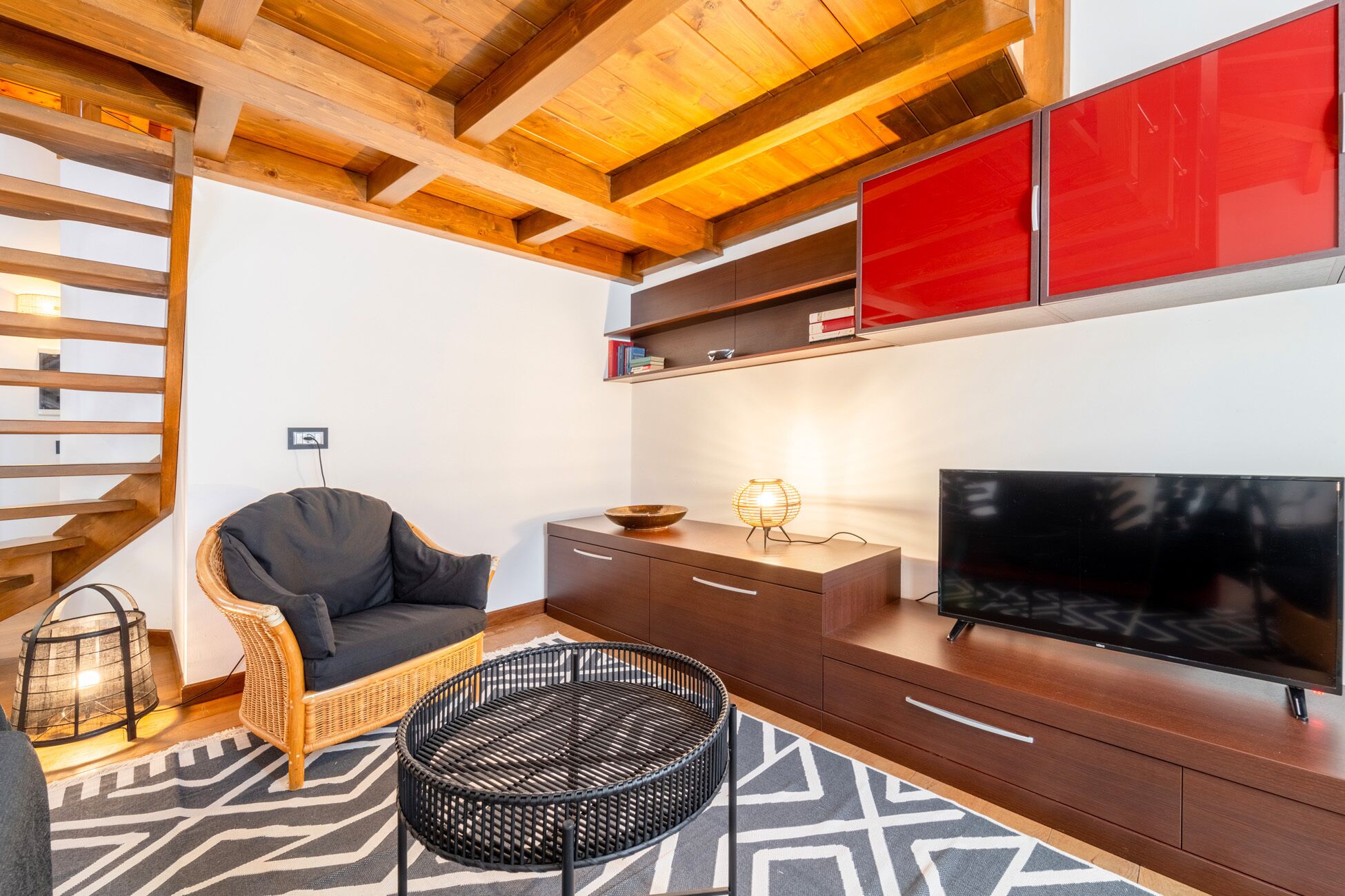 Una sala de estar moderna con techo de madera, televisor negro, silla de mimbre, mesa redonda negra y armarios rojos.