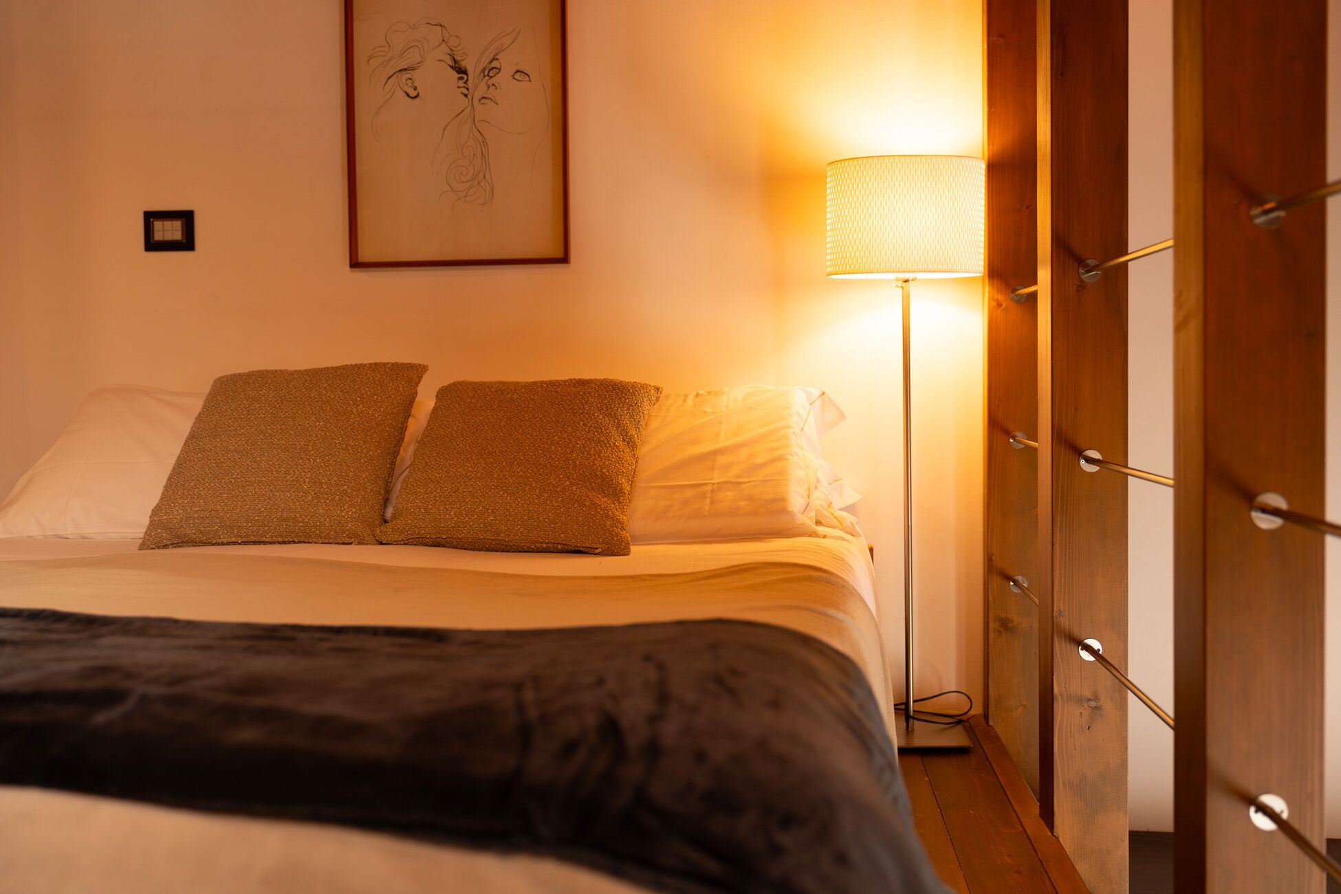 Un letto ben rifatto con due cuscini e una coperta scura. Una lampada da terra è posizionata a destra del letto, e un quadro incorniciato è appeso al muro sopra di esso.