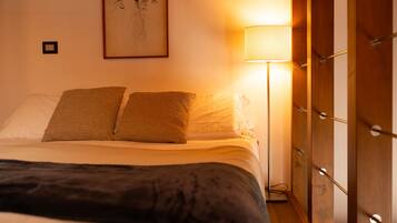 Un letto ben rifatto con due cuscini e una coperta scura. Una lampada da terra è posizionata a destra del letto, e un quadro incorniciato è appeso al muro sopra di esso.