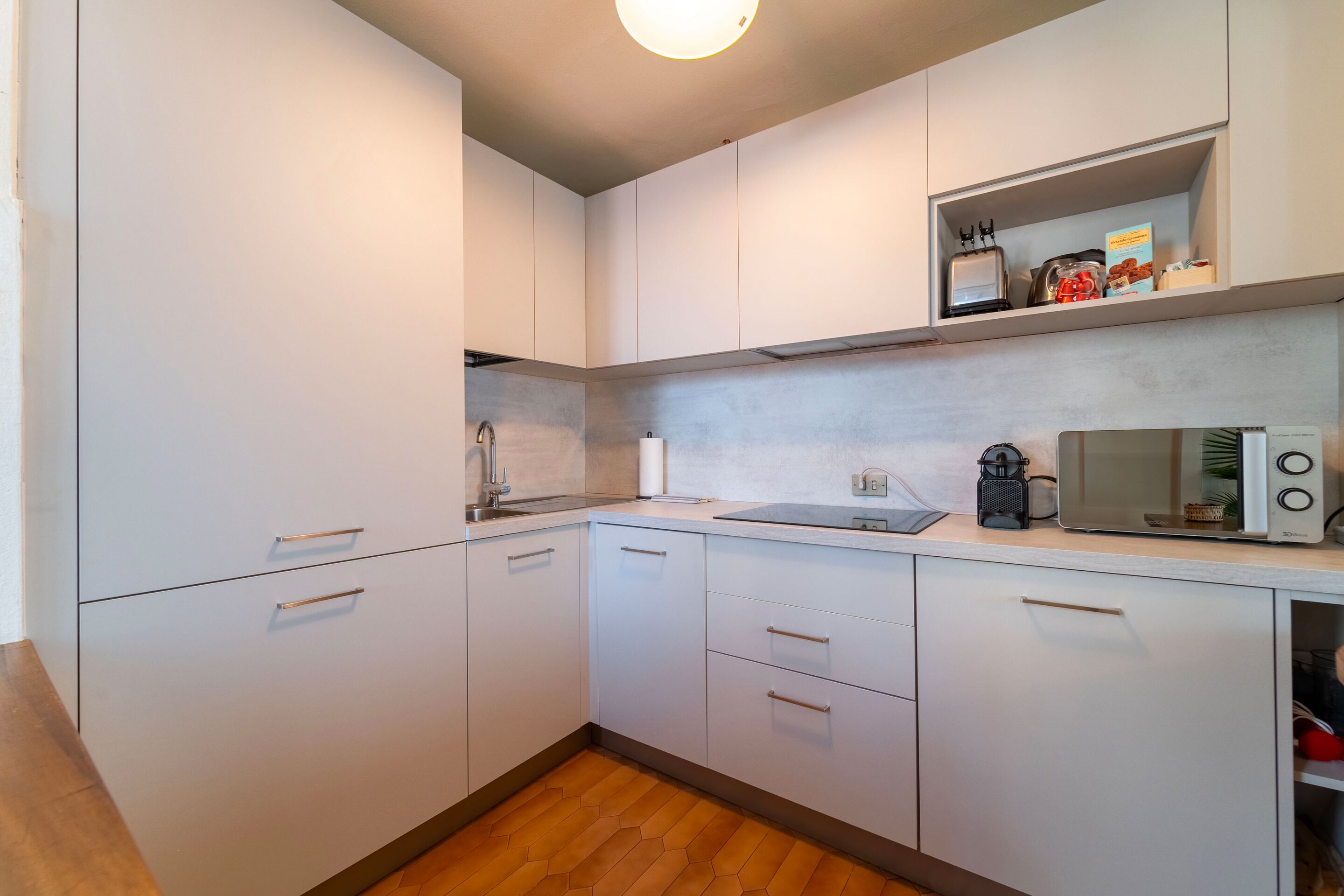 A modern kitchen with white cabinets, a microwave, a kettle, and a shelf with kitchen items.