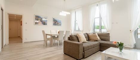 A modern living room with a brown sofa, white dining table, and chairs. There is a white coffee table with a vase of flowers.