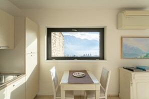 A compact kitchen with a window overlooking mountains, a dining table with a placemat, and a painting on the wall. - Zinnia Lakeview Apartment (Bellagio)