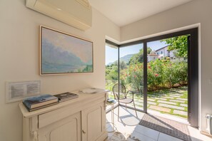 A room with a large window, a painting, a small table with books, and a white cabinet. - Zinnia Lakeview Apartment (Bellagio)