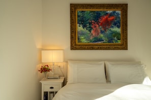 A neatly made bed with white linens, a bedside table with a lamp and a vase of flowers, and a framed painting on the wall. - Zinnia Lakeview Apartment (Bellagio)