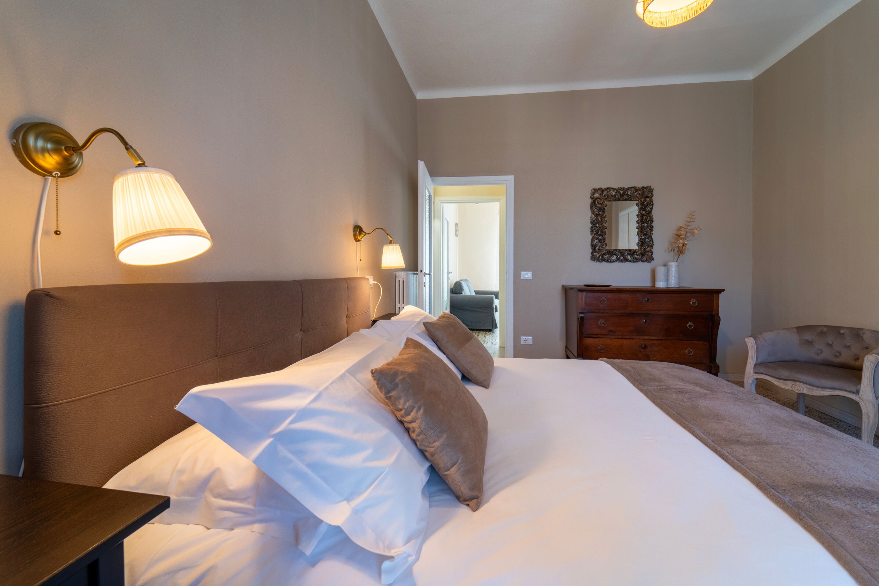 Un lit bien fait, avec du linge de lit blanc et des oreillers marron, une table de chevet en bois, une commode et deux lampes fixées au mur.