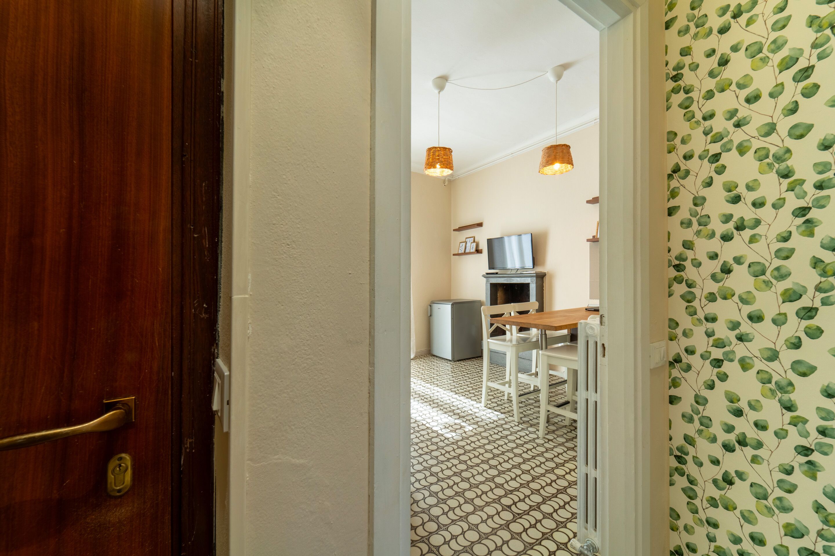 Un pasillo con puerta de madera, un radiador, una mesita con una silla y un televisor sobre un mueble bajo.