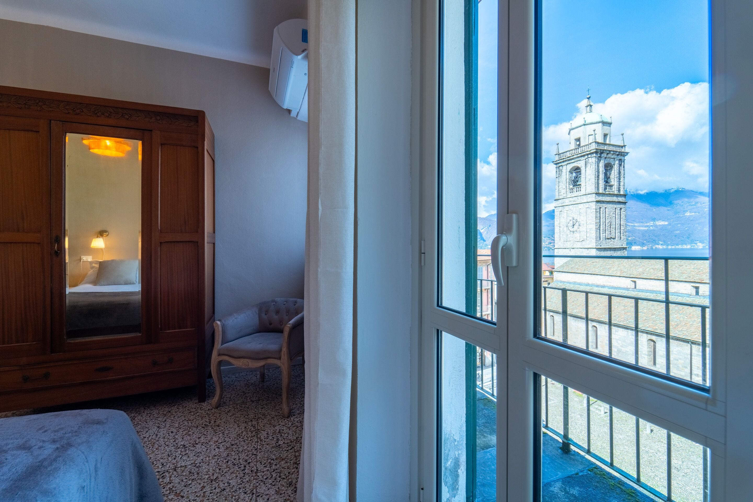 A room with a large wooden wardrobe, a chair, and a bed. A window reveals a clock tower and a mountainous landscape.