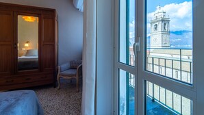 Une pièce comprenant une grande armoire en bois, une chaise et un lit. Une fenêtre donne sur un beffroi et un paysage montagneux.