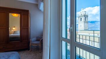 A room with a large wooden wardrobe, a chair, and a bed. A window reveals a clock tower and a mountainous landscape.
