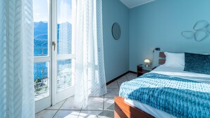 Une chambre avec une grande fenêtre, un lit recouvert d’une couverture bleue, un élément de décoration circulaire accroché au mur et une table de chevet avec une lampe.