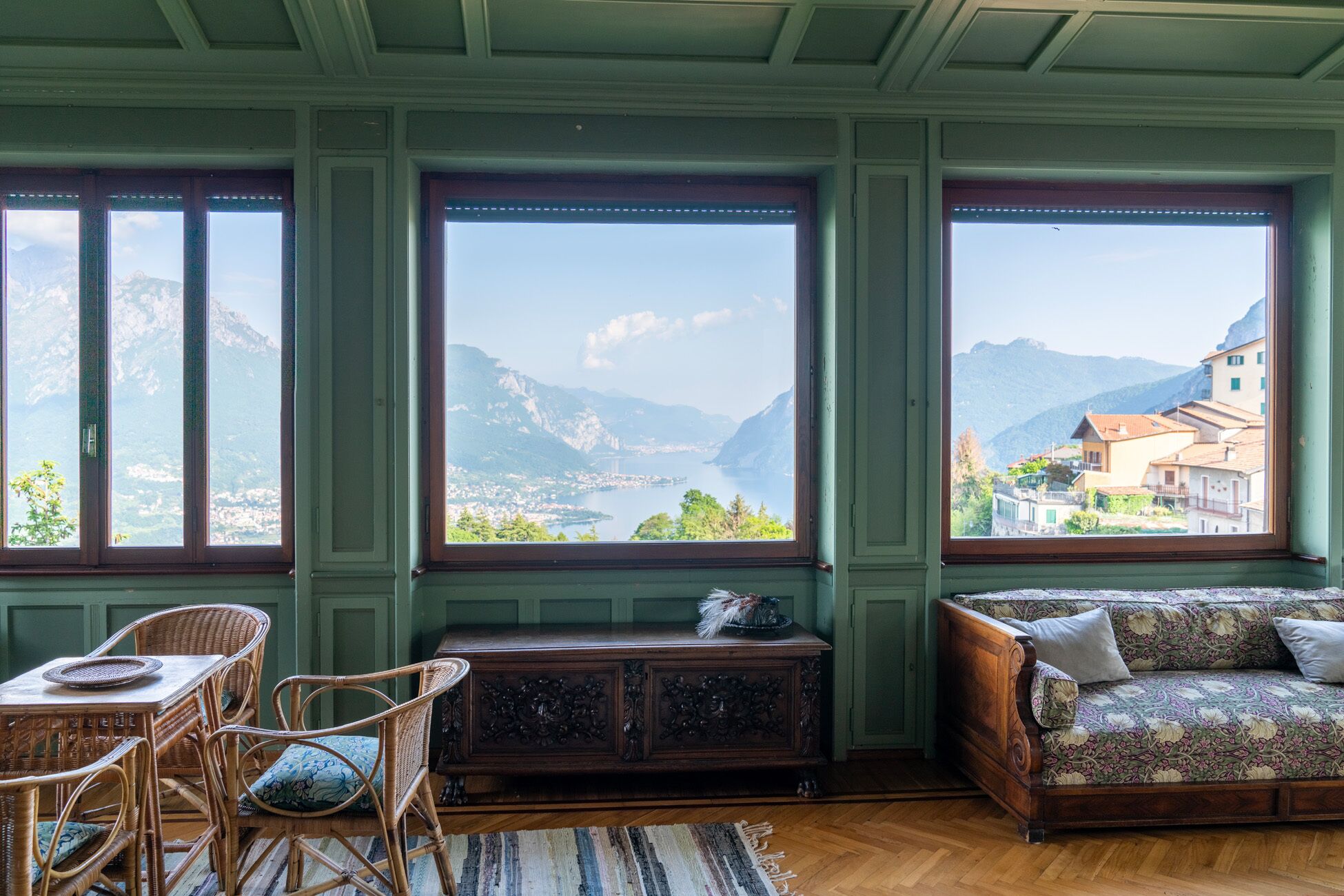 Une pièce avec vue sur les montagnes et un lac, un meuble en bois, des meubles en osier et un canapé à motifs.